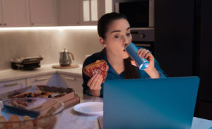 Young woman eating sandwich and drinking soda while using laptop